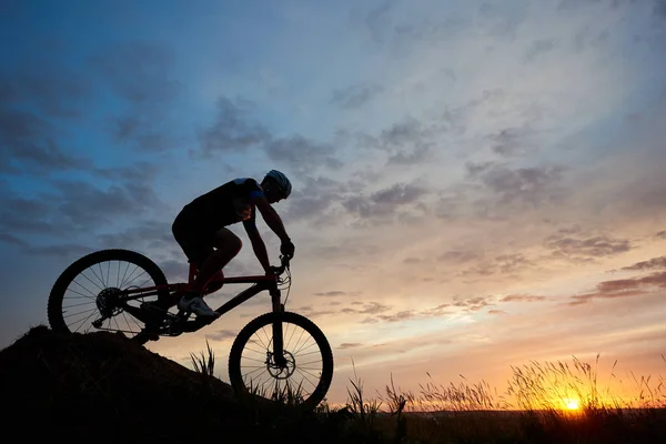 一个自行车的剪影戴着头盔和运动衫骑在山上的自行车下山下 黄昏的天空下的太阳日落 — 图库照片