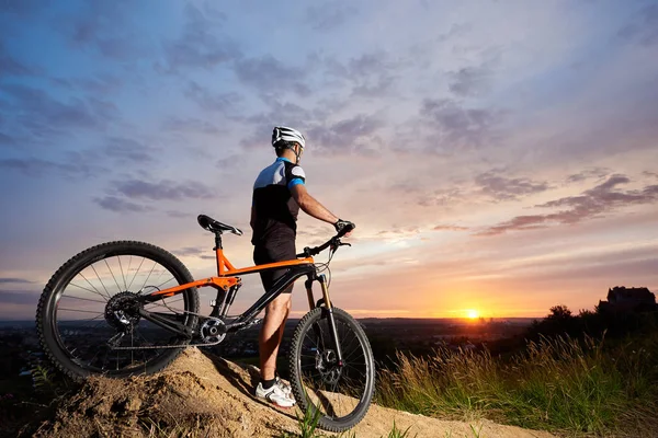 一个骑着山地自行车站在夕阳背景下的山上 坐着的太阳和一个美丽的蓝天下的城市的人后视图 — 图库照片