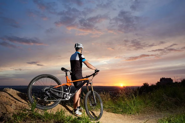 骑自行车的男性在头盔和运动服装的背面观看的背景下日落从高度看山在距离 迷人的天空和明亮的太阳 — 图库照片