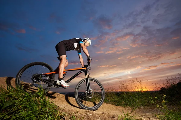 戴着头盔和运动服的家伙骑着一辆山地自行车下山 傍晚时分天空下着太阳日落 — 图库照片