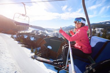 Genç, çekici bir kadın, kasklı, gözlüklü ve sıcak elbiseli gülümseyen bir kayakçı. Teleferikle, havada, baş parmak işaretini gösteriyor.