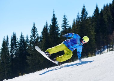Snowboardcu, gözlüklü ve miğferli bir adam, kar kaykayı sürerken hızlı, dik, karlı dağ yamaçlarında, güneşli kış günlerinde mavi gökyüzü ve ladin ağaçlarının arka planında.