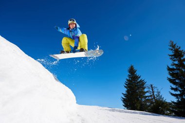 Snowboardcu, gözlüklü ve miğferli bir adam, kar kaykayı sürerken hızlı, dik, karlı dağ yamaçlarında, güneşli kış günlerinde mavi gökyüzü ve ladin ağaçlarının arka planında.