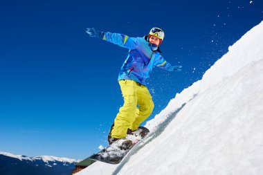 Snowboardcu, gözlüklü ve miğferli bir adam, kar kaykayı sürerken hızlı, dik, karlı dağ yamaçlarında, güneşli kış günlerinde mavi gökyüzü ve ladin ağaçlarının arka planında.