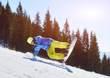 Snowboardcu, gözlüklü ve miğferli bir adam, kar kaykayı sürerken hızlı, dik, karlı dağ yamaçlarında, güneşli kış günlerinde mavi gökyüzü ve ladin ağaçlarının arka planında.