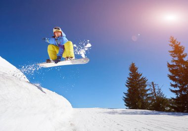 Snowboardcu, gözlüklü ve miğferli bir adam, kar kaykayı sürerken hızlı, dik, karlı dağ yamaçlarında, güneşli kış günlerinde mavi gökyüzü ve ladin ağaçlarının arka planında.