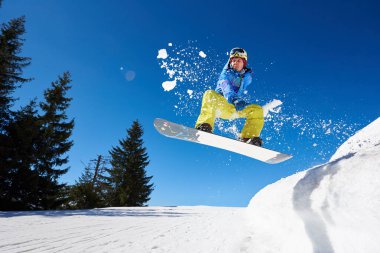 Snowboardcu, gözlüklü ve miğferli bir adam, kar kaykayı sürerken hızlı, dik, karlı dağ yamaçlarında, güneşli kış günlerinde mavi gökyüzü ve ladin ağaçlarının arka planında.