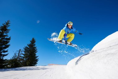 Snowboardcu, gözlüklü ve miğferli bir adam, kar kaykayı sürerken hızlı, dik, karlı dağ yamaçlarında, güneşli kış günlerinde mavi gökyüzü ve ladin ağaçlarının arka planında.