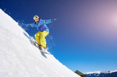Snowboardcu, gözlüklü ve miğferli bir adam, kar kaykayı sürerken hızlı, dik, karlı dağ yamaçlarında, güneşli kış günlerinde mavi gökyüzü ve ladin ağaçlarının arka planında.