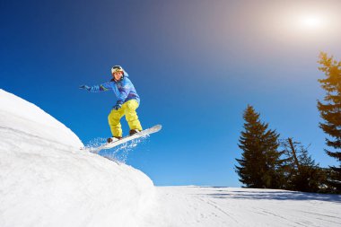 Snowboardcu, gözlüklü ve miğferli bir adam, kar kaykayı sürerken hızlı, dik, karlı dağ yamaçlarında, güneşli kış günlerinde mavi gökyüzü ve ladin ağaçlarının arka planında.