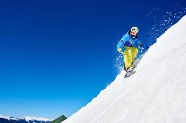 Snowboardcu, gözlüklü ve miğferli bir adam, kar kaykayı sürerken hızlı, dik, karlı dağ yamaçlarında, güneşli kış günlerinde mavi gökyüzü ve ladin ağaçlarının arka planında.