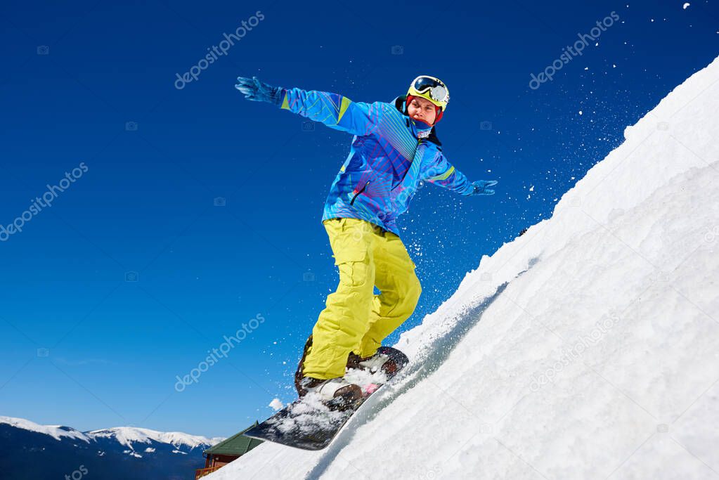 Snowboarder man in goggles and helmet riding snowboard fast down steep snowy mountain slope on background of blue sky and spruce trees on sunny winter day