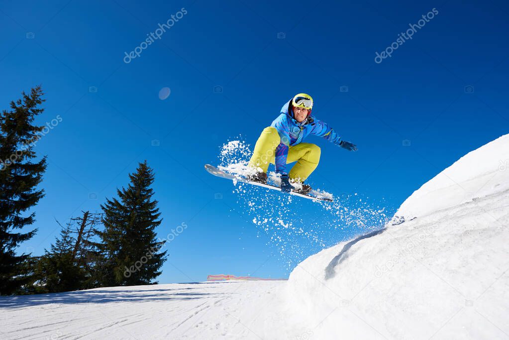 Snowboarder man in goggles and helmet riding snowboard fast down steep snowy mountain slope on background of blue sky and spruce trees on sunny winter day