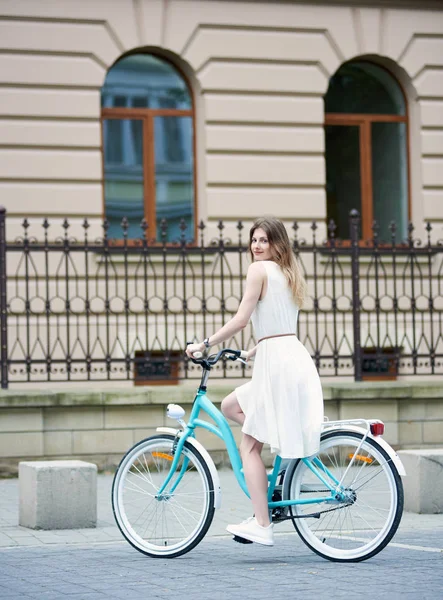 街の通りで青い自転車に乗っている薄い白いドレスと白いスニーカーの若い女性のリアビュー — ストック写真