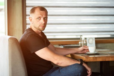 Yakışıklı, düşünceli, sportif bir adam elinde bir fincan kahve tutuyor, kafenin pencere kenarında oturuyor, siyah tişört ve koyu mavi kot pantolon giyiyor.