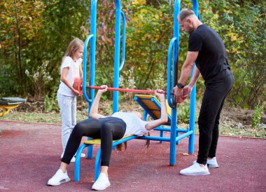 Genç bir aile açık hava spor salonunda aktif zaman geçiriyor, karısı uzanıyor ve her iki tarafta duran kocası ve kızının yardımıyla kırmızı halteri kaldırıyor.