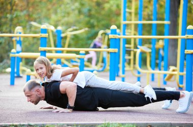 Babası açık havada, sırtında kızıyla şınav çekiyor. Sağlıklı bir yaşam tarzı, aile, yan görüş, arka planda açık spor salonu.