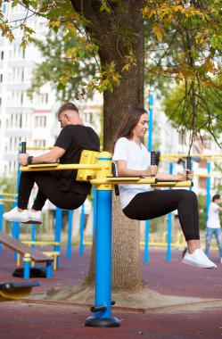 Karı koca karın kaslarını çalıştırıyorlar. Spor salonunda bacaklarını arka arkaya kaldırıyorlar. Sağlıklı yaşam tarzı, arka planda büyük bir ağaç.
