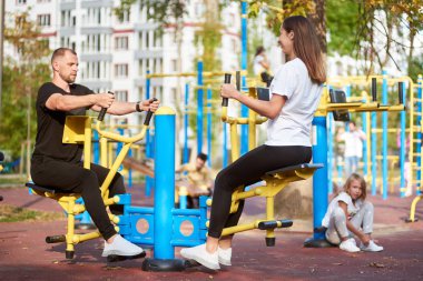 Anne ve baba spor barlarında kollarını ve bacaklarını yüz yüze çalıştırıyor. Kızları arka planda, güneşli havada.