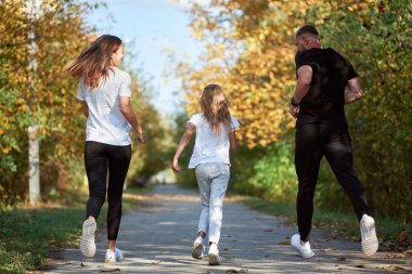 Anne, baba ve kızın spor giyim kuşamlarının arka planı sonbahar parkındaki sokakta, aktif ailede, sıcak ve güneşli havada koşuyor.