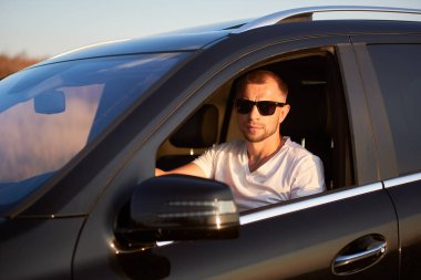 Beyaz tişörtlü, kendine güvenen ciddi bir adam siyah gözlüklü siyah arabasında oturuyor, pencereden dışarı bakıyor, yan görünüş, yakın çekim.