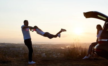 Bir babanın silueti kızını elinden tutuyor, annesi tepenin üstünde arabanın bagajında oturuyor. Arka planda batan güneşin güzel manzarası, yan manzara