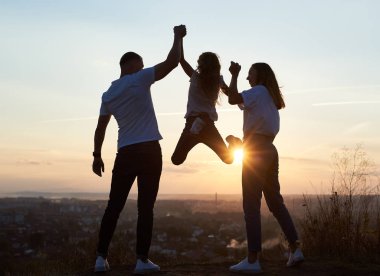 Anne ve kızının silueti şehrin dışında gün batımında güzel bir şehir manzarası olan tepenin üstünde, baba ve anne kızın elini tutuyor ve kız asılı duruyor.