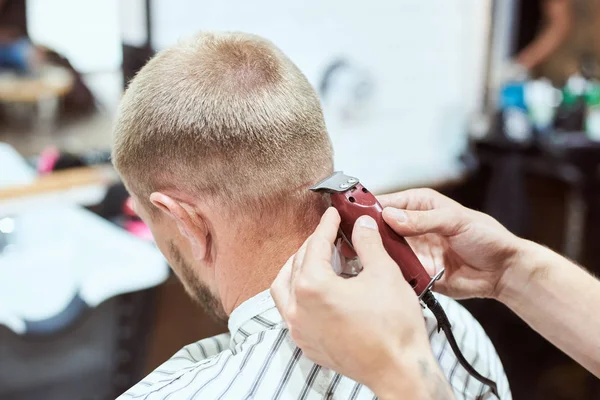 Dark blonde man at barber shop is having his nape shaved with a trimmer, hairdresser is holding a red tool, stripe cape on