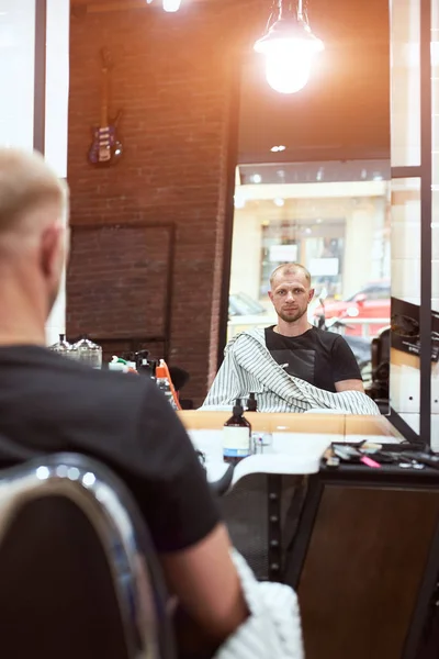 年轻英俊的留着胡子的男人坐在理发店的椅子上 看着镜子里自己的倒影 — 图库照片