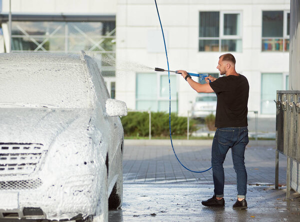 Full length view of man in dark clothes is washing his car holding a hose with foam outdoors, side view