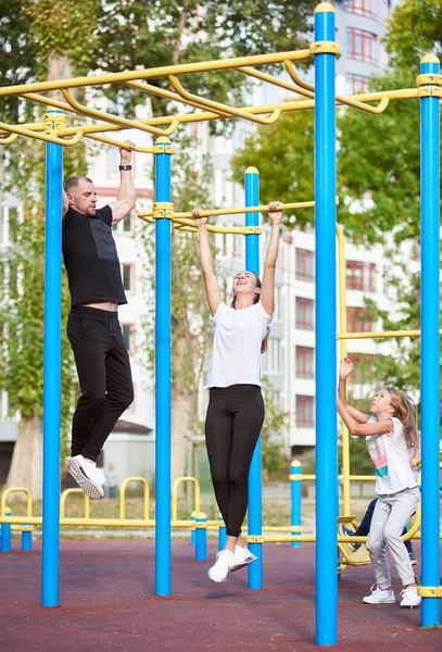 Beautiful family exercising at the outdoor gym, man and woman doing chin ups on the sport bars, residential building on the background