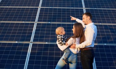 Güneşli bir günde, fotovoltaik güneş panelinin yanında bir bebeği bir arada tutan güzel, genç bir ailenin arka planı, fotokopi alanı, modern aile kavramı.