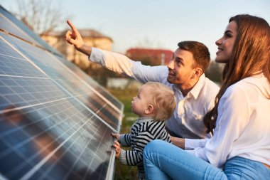 Adam sıcak bir günde evin yakınındaki arsadaki güneş panellerini ailesine gösteriyor. Çocuklu genç bir kadın ve güneş ışınlarındaki bir adam güneş panellerine bakıyor..