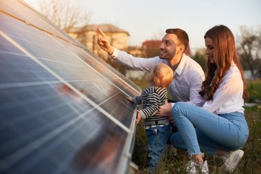 Side view shot of a young modern family with a little baby boy getting acquainted with solar panel on a sunny day, green alternative energy concept clipart