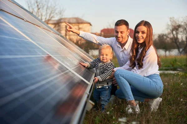 一个年轻的三口之家蹲在光伏太阳能电池板旁 微笑着看着相机 这是一个光明的未来的概念 — 图库照片