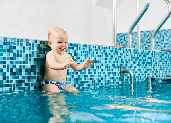 Cute playful baby boy is sitting on a silver pipe in the swimming pool preparing to jump in water to his mom\'s hands. Concept of healthy chindhood