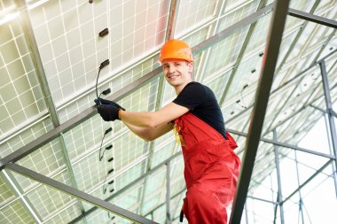 Erkek mühendis kaliteli güneş panellerini inceliyor. Turuncu üniformalı ve kasklı bir işçi, kameraya gülümsüyor. Fotovoltaik güç tedarik sistemleri. Yeşil enerji konsept tasarımı.