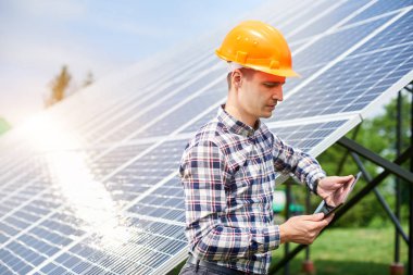 Kasklı erkek mühendis güneş panelleri istasyonunun yakınındaki elinde bulunan güneşten yansıyan tablete bakıyor. Ev yapımı. Yeşil ekolojik enerji üretimi