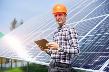 Güneş panelleri istasyonunun yanında elinde tabletle güneşli bir günde kask takan bir adamın portresi. Yeşil ekolojik enerji üretimi.
