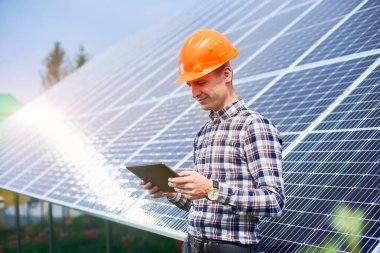 Güneş pili istasyonunun yakınında elinde tablet olan kasklı bir adam, içinde güneşin yansıması olan bir adam. Ev yapımı. Teknoloji konsepti. Yeşil ekolojik enerji üretimi
