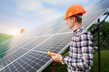 Güneş panellerinin yanında elinde bir sensör bulunan erkek işçi mavi gökyüzünün altındaki güneş ışınlarını yansıtıyor. Yeşil arka planda düz güneş. Elektrik santrali. Ev yapımı.