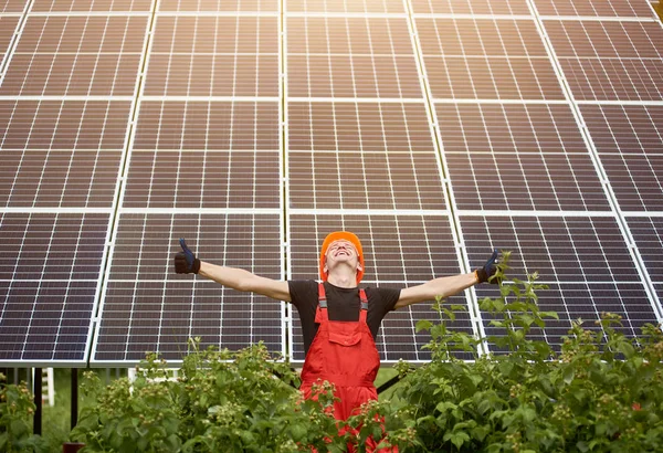 Happy worker raising his hands, showing thumbs signs like on a background of photovoltaic panels solar station. Man in orange uniform. Science solar energy. Renewable energy.