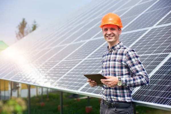 Güneş panelleri istasyonunun yanında elinde tabletle güneşli bir günde kask takan mutlu erkek işçinin portresi. Yeşil ekolojik enerji üretimi.