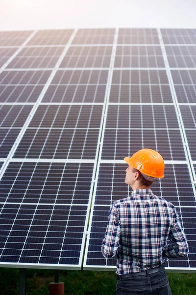 Engineer in an orange helmet stands with his back to the camera against the background of a large plantation of solar panels. Home construction. Green ecological power energy generation.