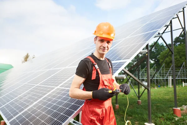 Worker in orange uniform and black gloves with equipment in hands working at solar station, looking to the camera. On background of solar panels against of greenery and blue sky. Home construction