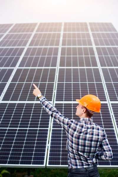 Back view of engineer in orange helmet pointing of a large station of solar panels. Home construction. Green ecological power energy generation.