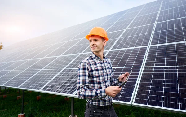 Portrait of man in a helmet with a tablet in his hands standing near the solar panels station. Green ecological power energy generation. Solar station development concept. Home construction.