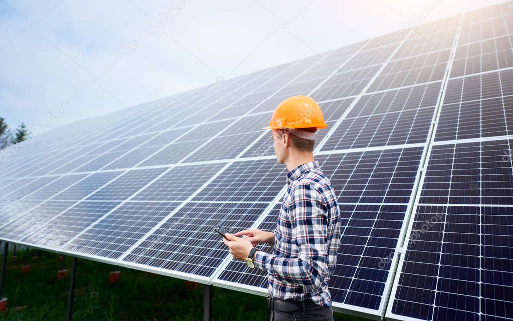 Engineer in helmet with tablet in hands standing near solar panels on green plantation. Concept ecology protection. Man worker. Plant industry.