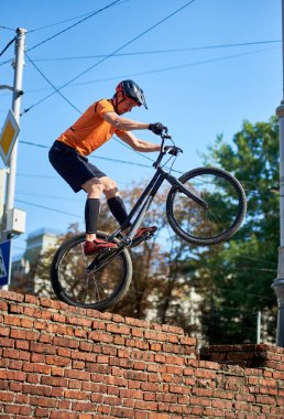 Side view of guy in black shorts stands on one wheel of a sports bike. Serious man cycling in city on nature background. Concept of sport.