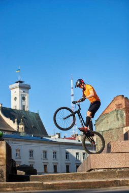 Side view of a cyclist standing on a black bike. Focused athlete preparing doing trick in city. Athlete holding a mountain bike and standing on one wheel. Concept of keeping balance.